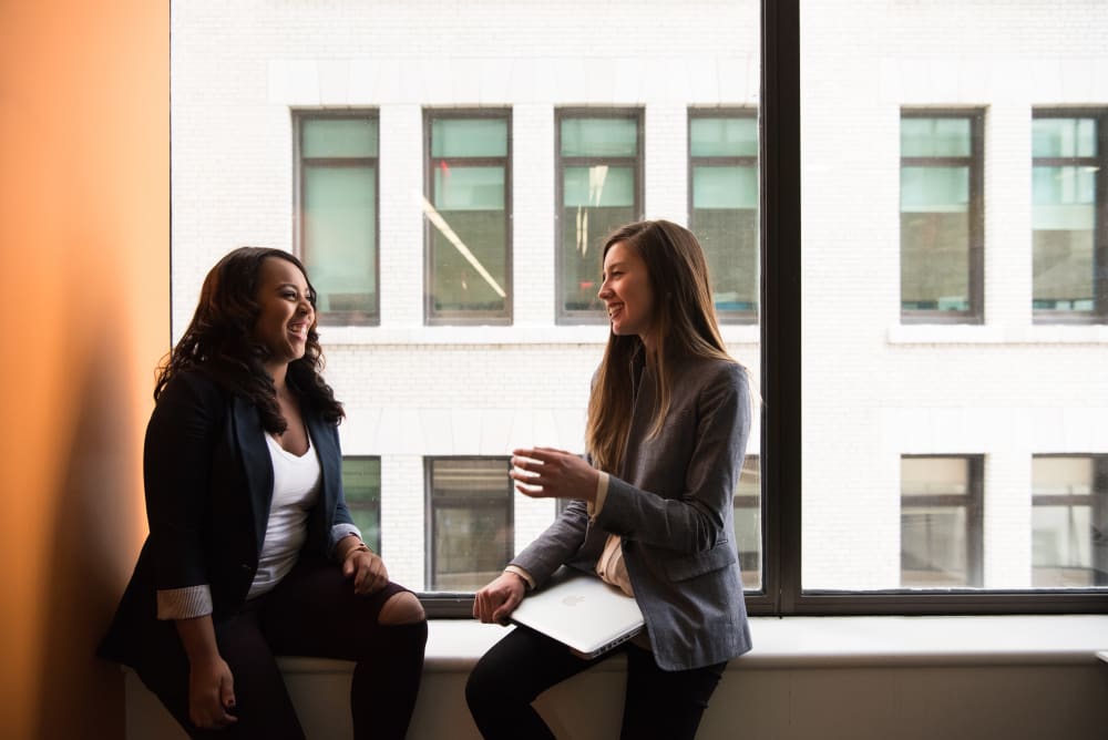 Two business women, talking to one another and smiling, sitting by a window within an inner city office