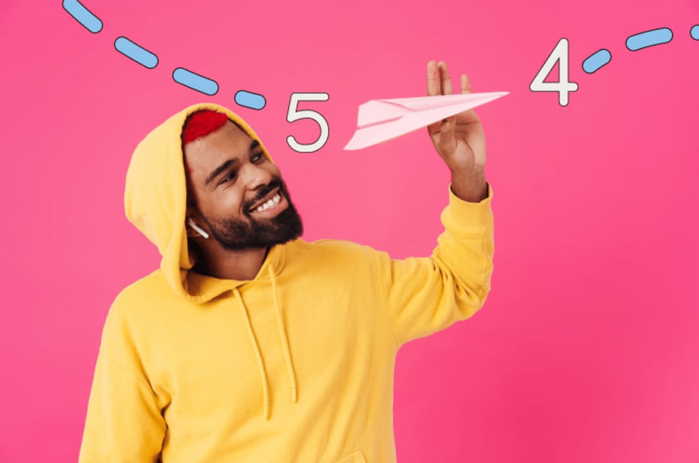 Man in a yellow hoodie holding a paper airplane with a pink background