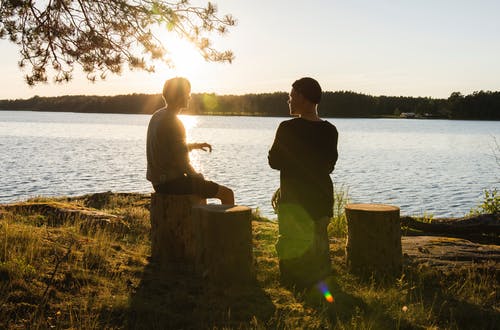 Man and a woman sitting on tree stumps in front of a lake at sunset