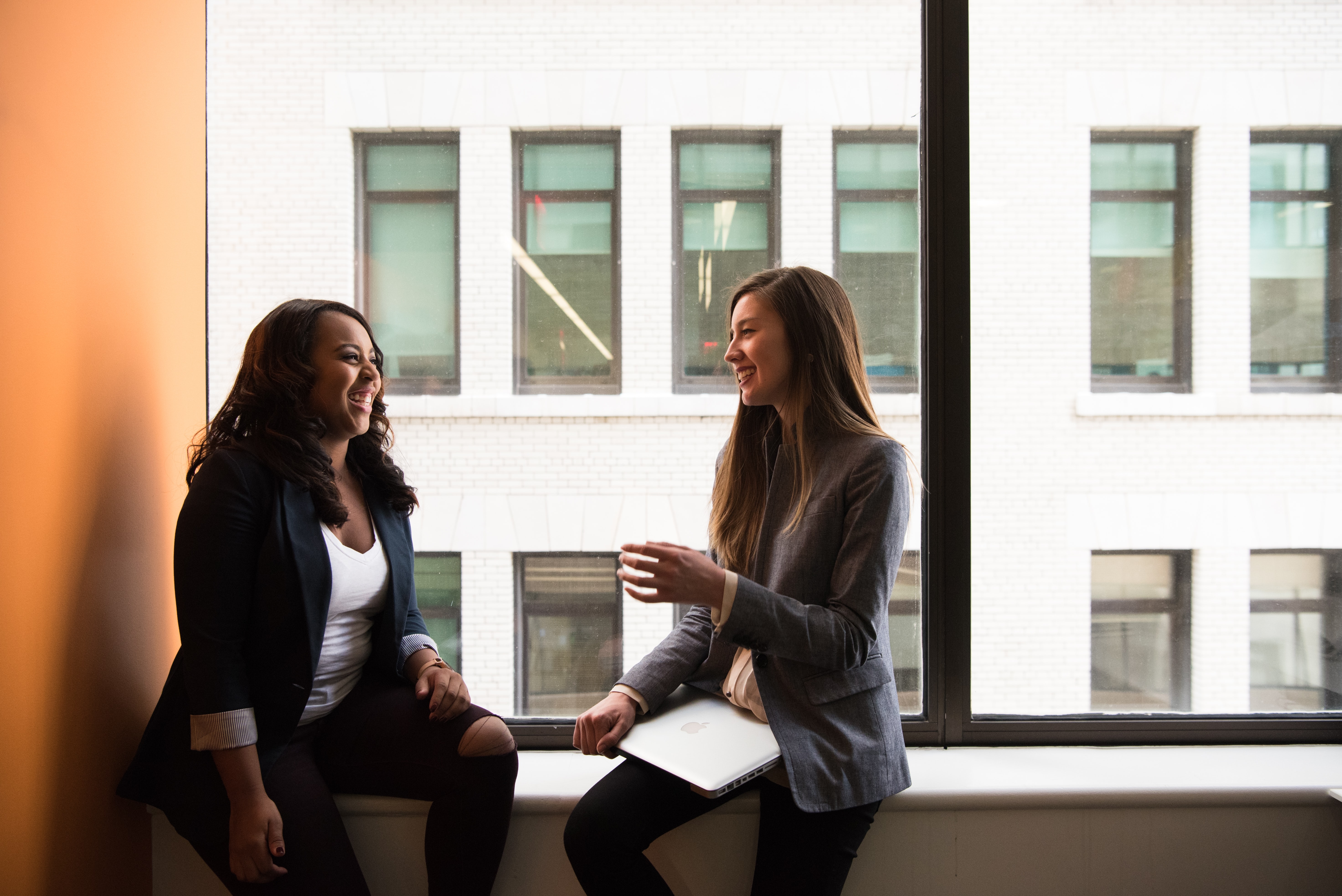 Two business women, talking to one another and smiling, sitting by a window within an inner city office.