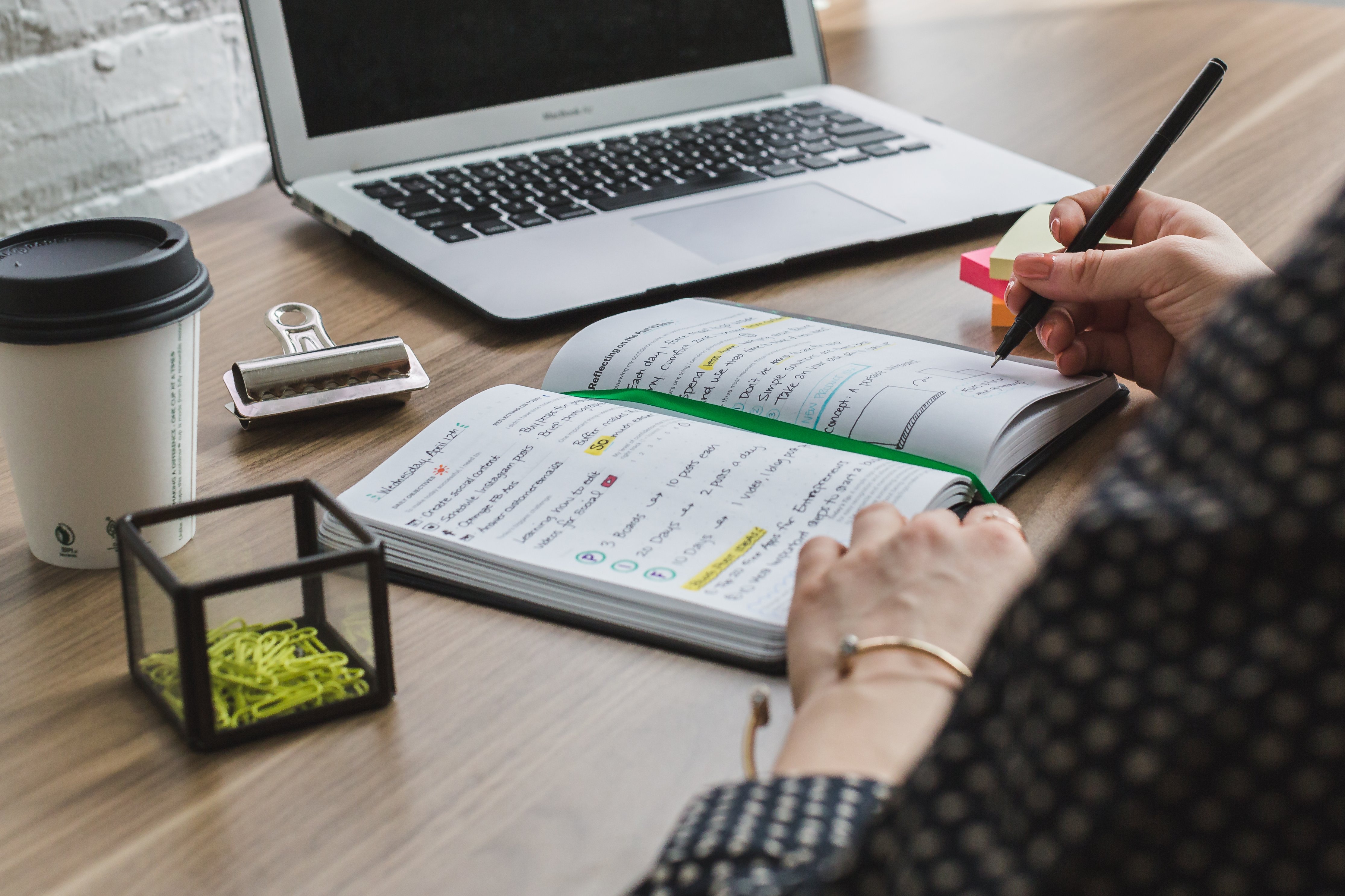 Woman writing in her diary at a work desk in front of her laptop and an empty coffee mug